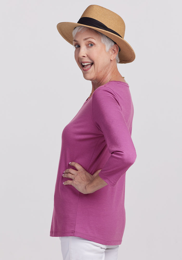 Smiling older woman in a straw hat and white pants models the Woolx Jenny 3/4 Sleeve Crew Neck in Purple Wine, posing sideways with one hand on her hip against a light background.