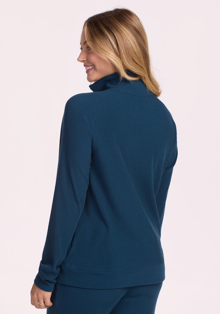 A woman with light brown hair wears the Woolx Quinn Ribbed 1/4 Zip in Majolica Blue with matching pants, standing against a plain light background and smiling over her shoulder.