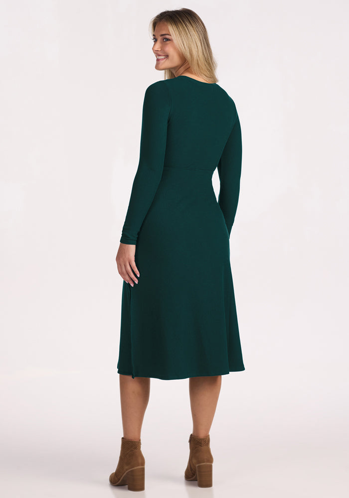 A woman stands facing away, turning slightly to smile at the camera. She wears the Woolx Caroline Ribbed Twirl Dress in Deep Timber and brown ankle boots, set against a plain white background.