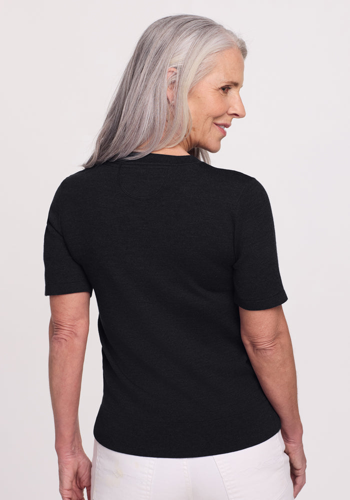 An older woman with long gray hair stands slightly turned to the side, wearing the Woolx Anna Crew Sweater in black and white pants against a plain light background.