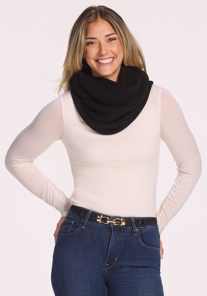 A woman with long blonde hair smiles while wearing the Woolx Noelle Infinity Scarf in black, styled with a white long-sleeve top, dark jeans, and a black belt with a gold buckle against a plain light background—a perfect winter accessory.