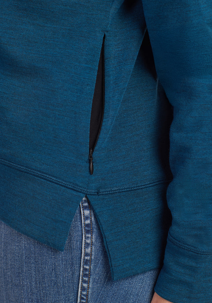 Close-up of a person wearing blue denim jeans and the Woolx Annie 3 Button Pullover in Majolica Blue, highlighting the partially unzipped side zipper pocket and lower side seam of the sweatshirt.