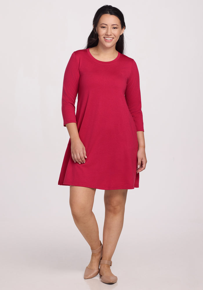 A woman with long dark hair smiles in the Woolx Rissa A Line Dress - Poppy Spark, a knee-length, bright red Merino wool dress with three-quarter sleeves and beige flats—ideal for fall/winter layering against a plain light background. 