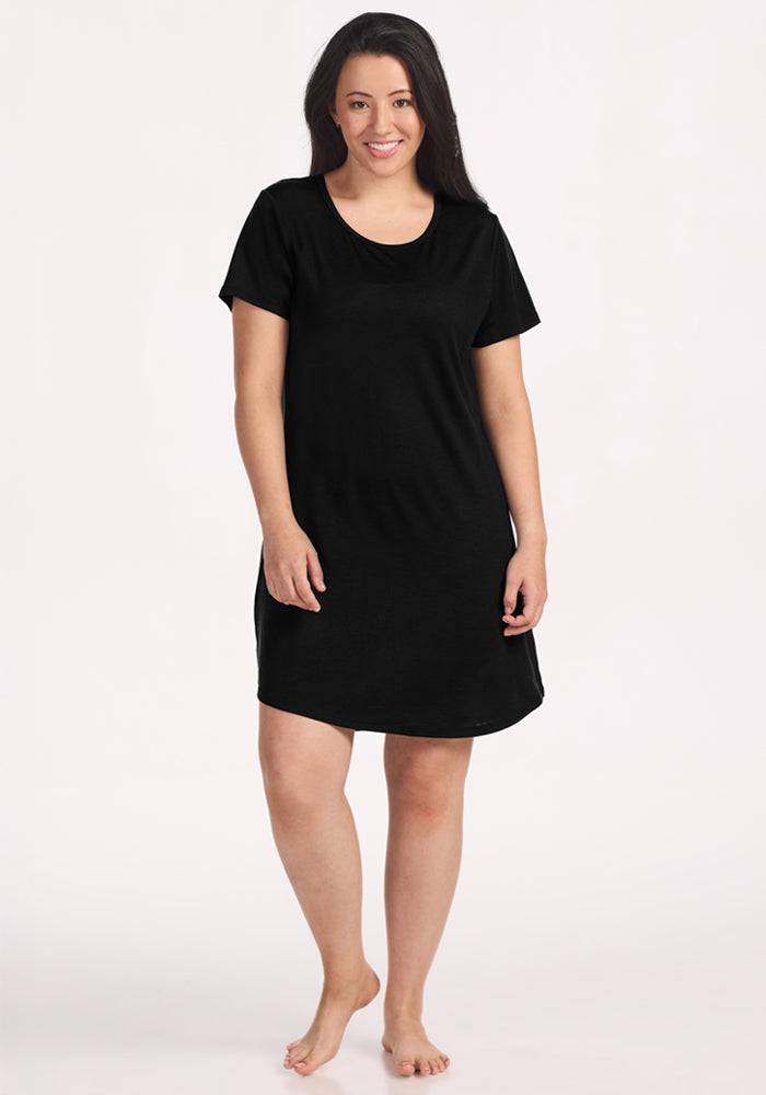 A barefoot woman with long dark hair smiles in the Woolx Desi Nightgown - Black, a simple Merino wool sleepwear piece, perfect for comfort and temperature regulation against a plain white background. 