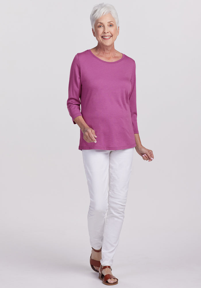 A smiling older woman with short gray hair wears the Woolx Jenny 3/4 Sleeve Crew Neck in Purple Wine, paired with white pants and brown sandals, standing against a plain light background.