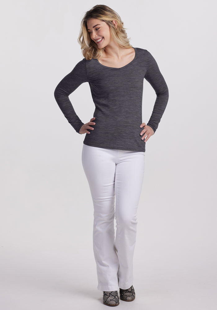 A blonde woman smiles against a white background, wearing the Woolx Layla V Neck Top in Cinder Melange with white pants and snakeskin-patterned shoes. She poses with hands on her hips, looking to the side.