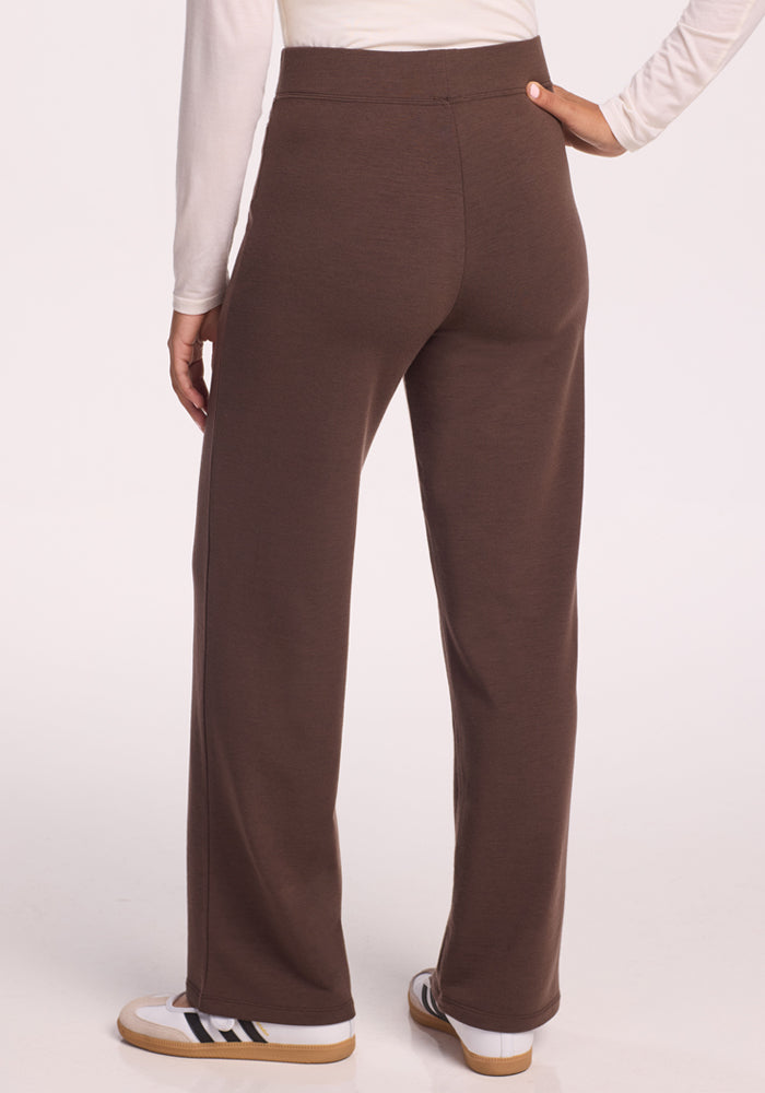 A petite woman is seen from the back, wearing Woolx Ellie Wide Leg Pants Petite in Bracken, a long-sleeved white top, and white sneakers with black and tan accents against a plain background.