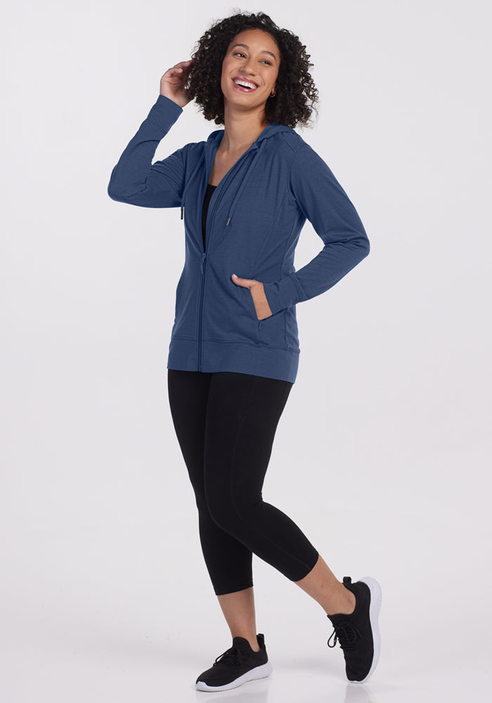 A smiling woman with curly hair models the Woolx Ryann Hoodie in Starry Night, paired with black leggings and sneakers. Posing on one leg against a white background, she highlights this travel essential’s style and comfort.