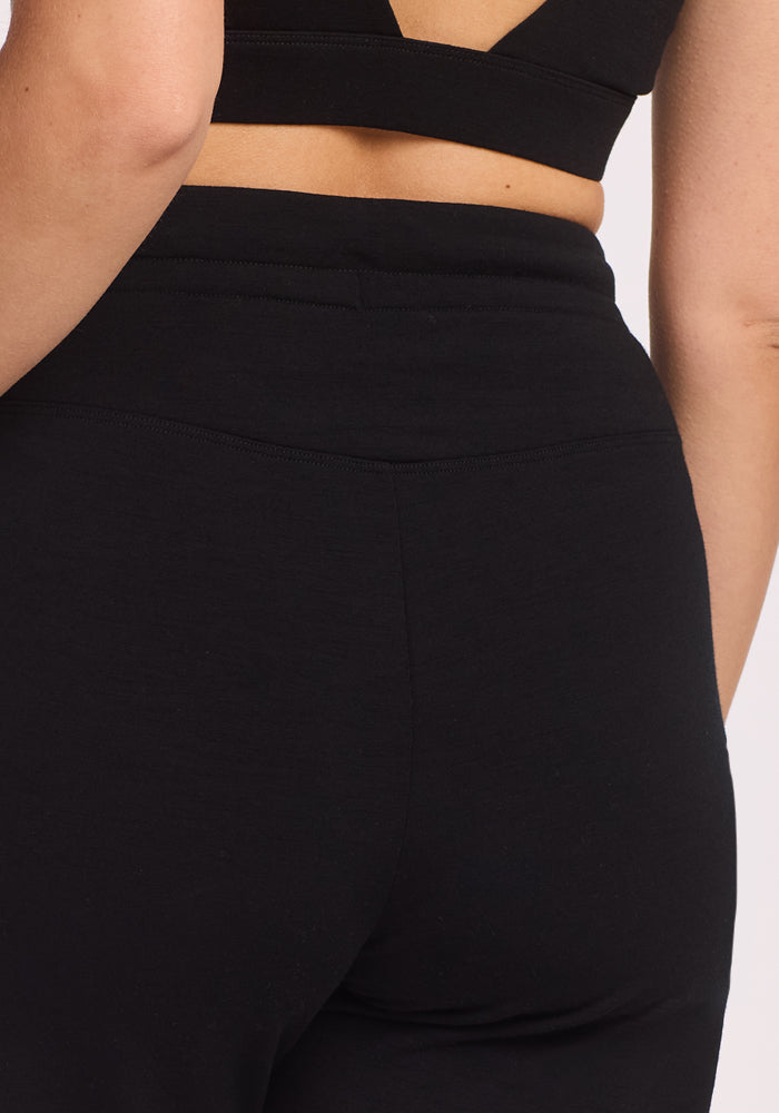 A close-up shows someone from behind wearing Woolx Willa Joggers Petite in black, paired with a black open-back top that reveals their lower back and waist.