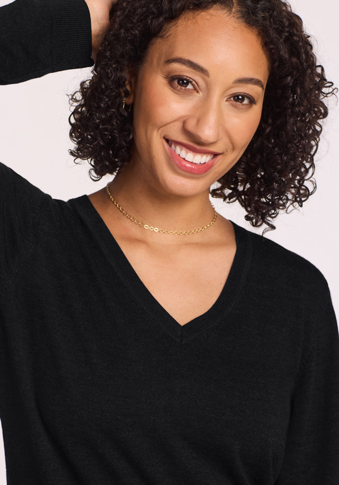 A smiling woman with curly hair poses with one arm raised against a light background, wearing the Woolx Francesca Sweater in Black and a gold chain necklace.