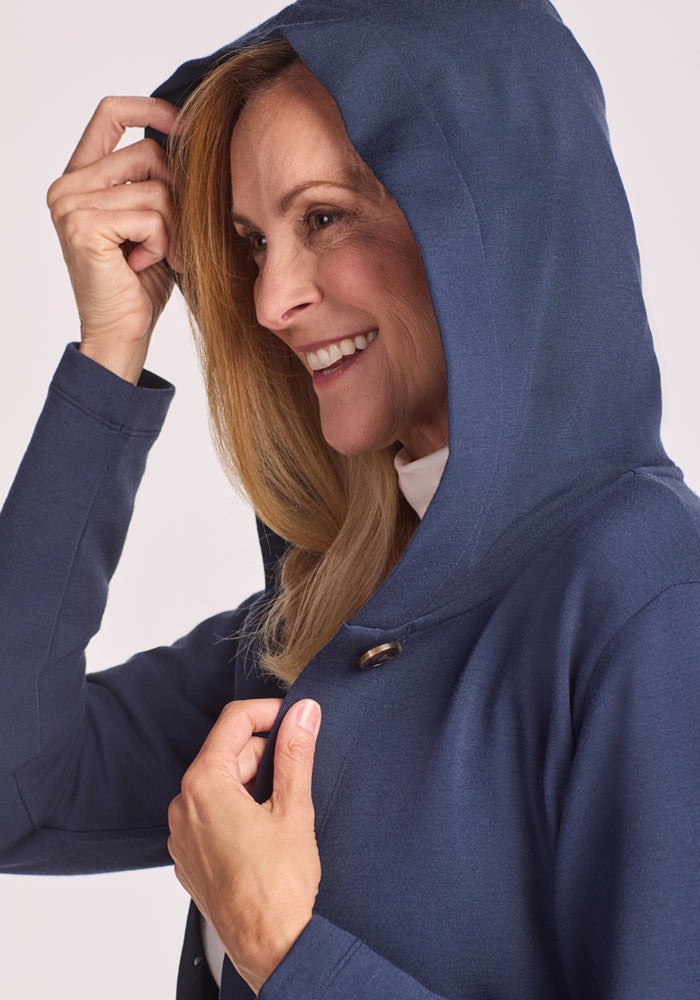 A smiling blonde woman wears the Woolx Edna Hooded Cardigan in Deep Navy, holding the hood with one hand and pulling it slightly forward. She faces left, looking happy and relaxed.