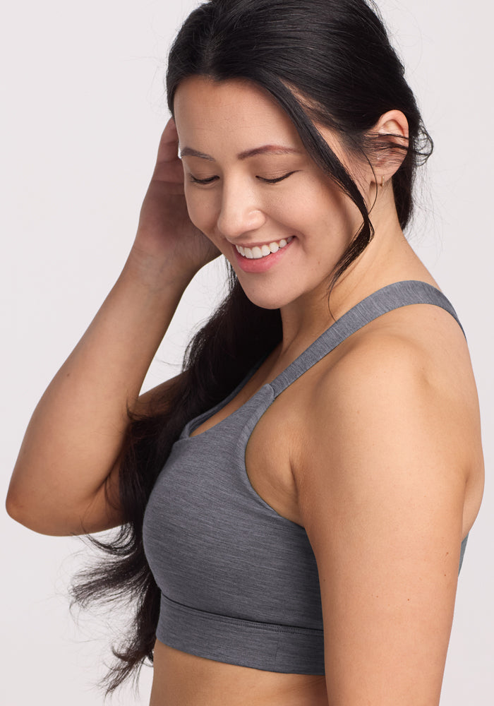 A woman with long dark hair smiles with her eyes closed, wearing the Woolx Livi Bra in Graphite Heather. She touches her hair and stands against a plain light background.