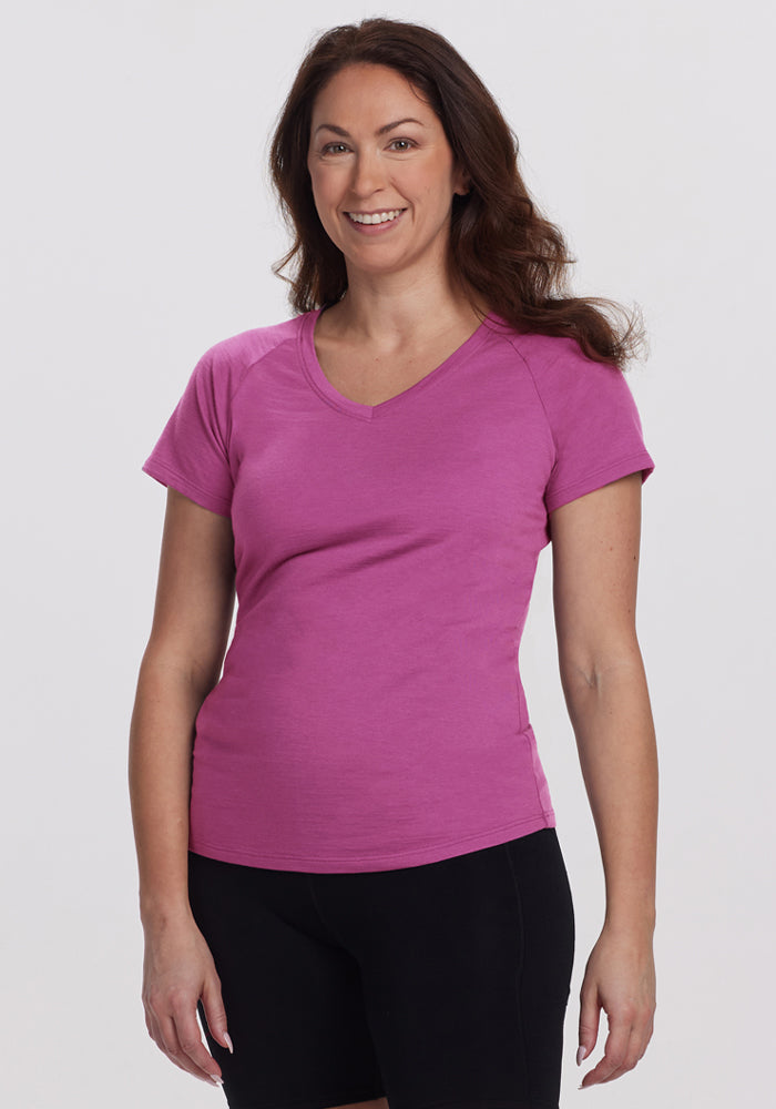 A woman with long brown hair, wearing the Woolx Mia V Neck in Purple Wine and black shorts, stands smiling against a plain light background. 