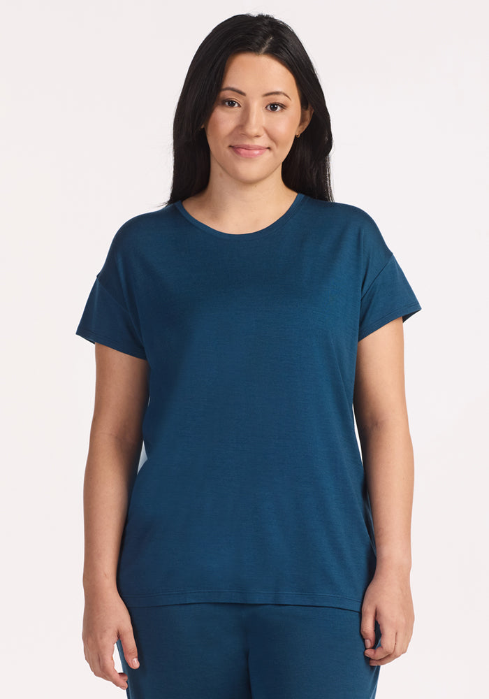 A woman with long dark hair faces the camera, smiling slightly. She wears the Woolx Billie Lounge Tee in Majolica Blue against a plain light background. 