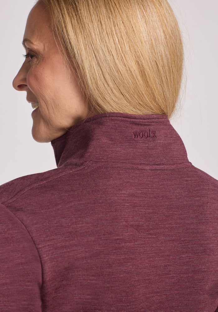 A woman with straight blonde hair is shown from the side and back, wearing the Woolx Finley Zip Up Sweatshirt in Grape Harvest Melange, featuring odor-resistant Merino wool and "woolx" embroidered on the collar.