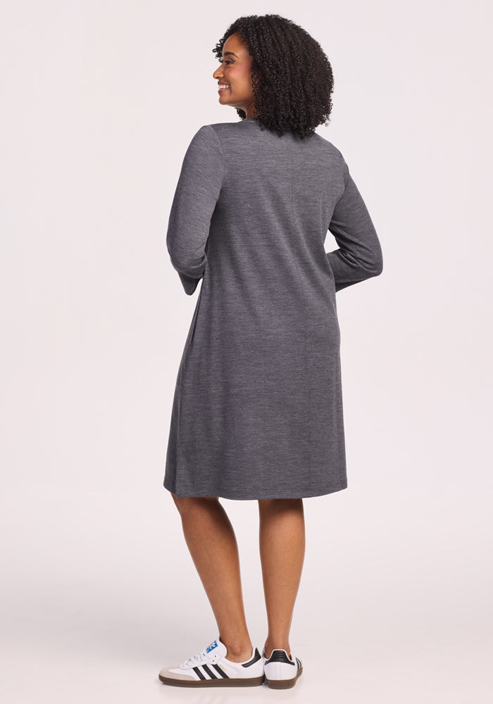 A woman with curly hair, wearing the Woolx Rissa A Line Dress in Cinder Melange and white Adidas sneakers with black stripes, stands smiling against a light background—ideal for fall and winter layering.