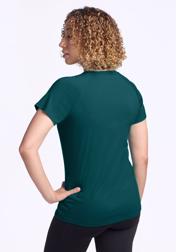 Someone with curly hair is standing with their back to the camera against a plain white background, wearing the Woolx Mia V Neck in Riverbank Green and black pants.