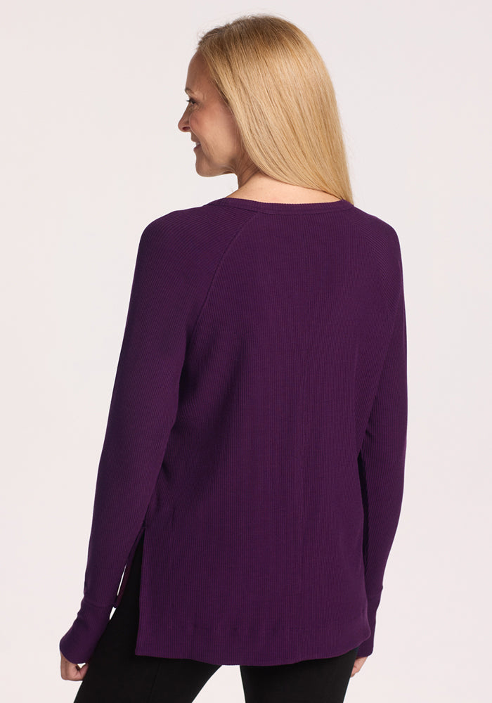A woman with straight blonde hair smiles while facing away from the camera, wearing Woolx's Ainsley Sweater in Majestic Purple and black pants against a light background.