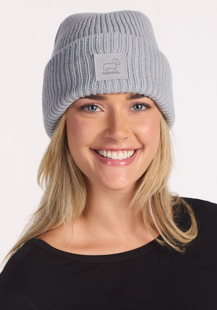 A smiling person with long blonde hair wears the Woolx Ria Chunky Beanie in Morning Fog, featuring a front logo patch, paired with a black shirt against a white background—the perfect winter accessory.