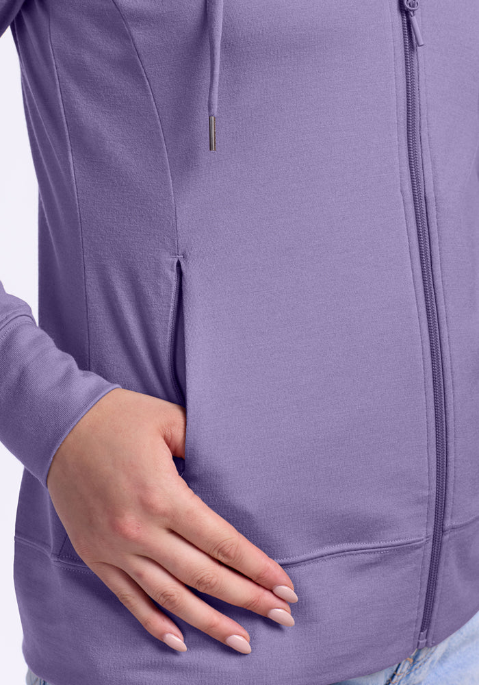 A person with light skin rests their hand on the pocket of the Woolx Ryann Hoodie in Montana Grape, revealing manicured nails and part of their blue jeans.