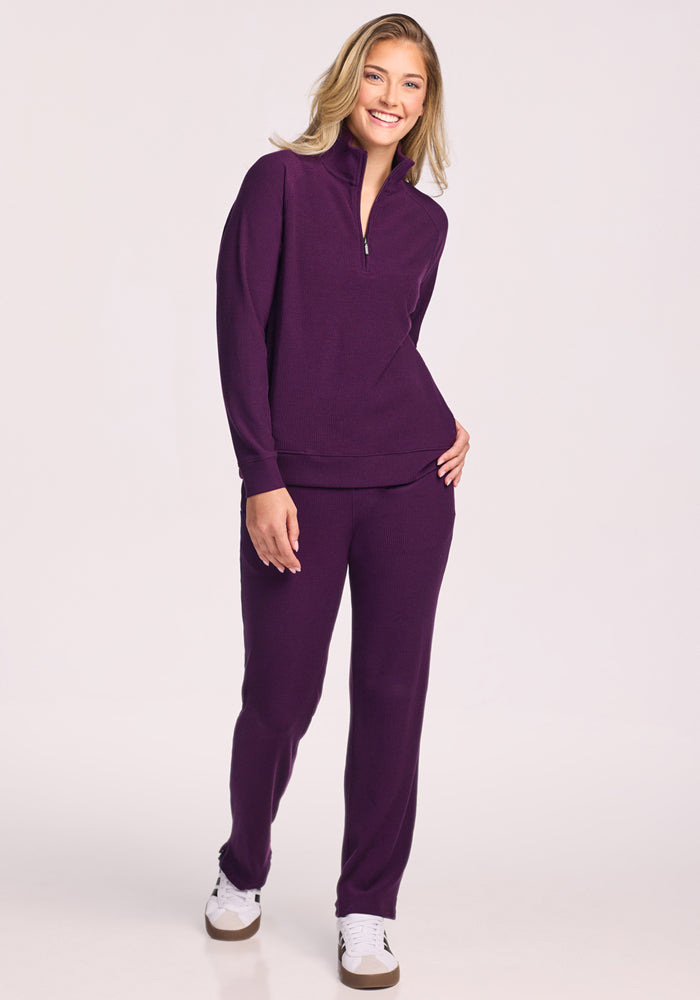 A smiling woman with blonde hair wears the Woolx Quinn Ribbed 1/4 Zip in Majestic Purple as a set with matching pants and white sneakers, standing against a plain light background.