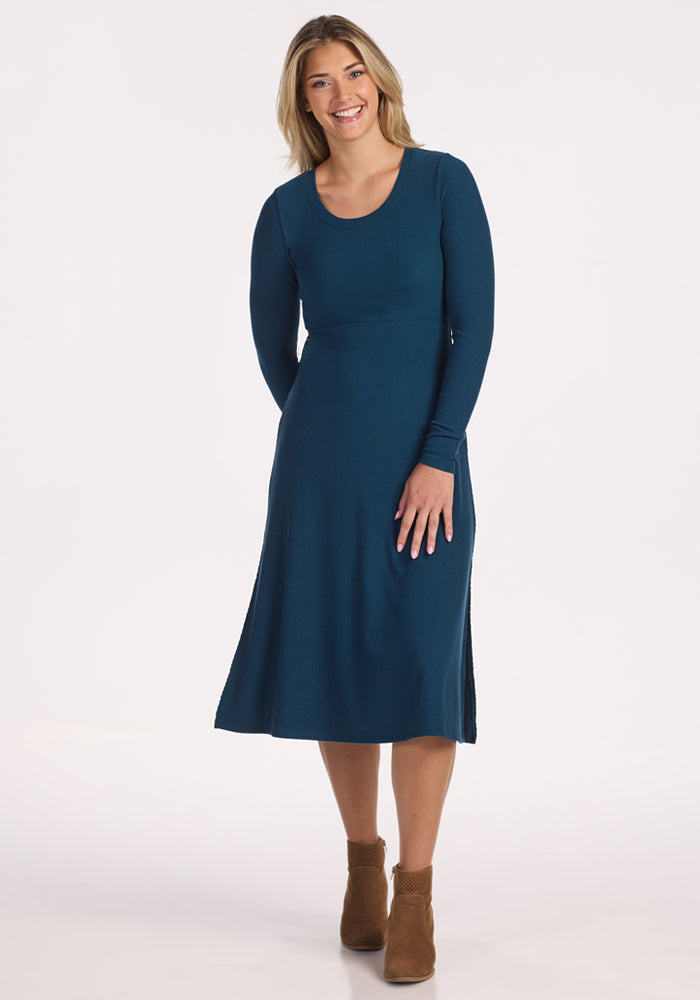 A woman with long blonde hair smiles against a white background, wearing the Woolx Caroline Ribbed Twirl Dress in Majolica Blue and tan ankle boots. 