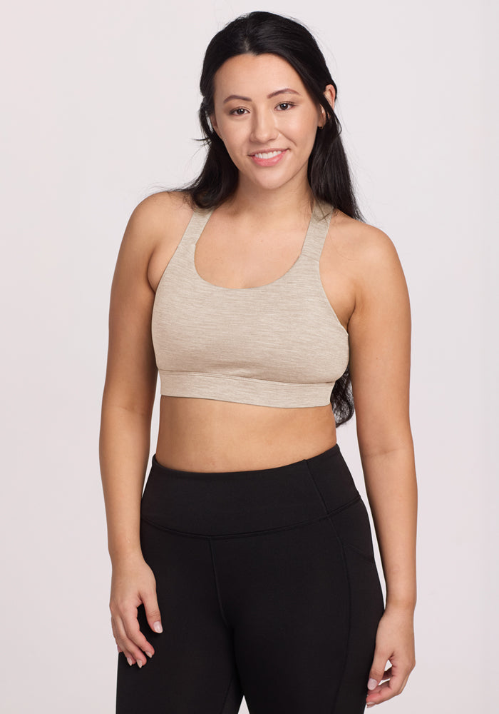 A woman with long dark hair smiles at the camera, wearing the Woolx Livi Bra in Cream Heather and black leggings, standing against a plain light background with her arms relaxed by her sides. 