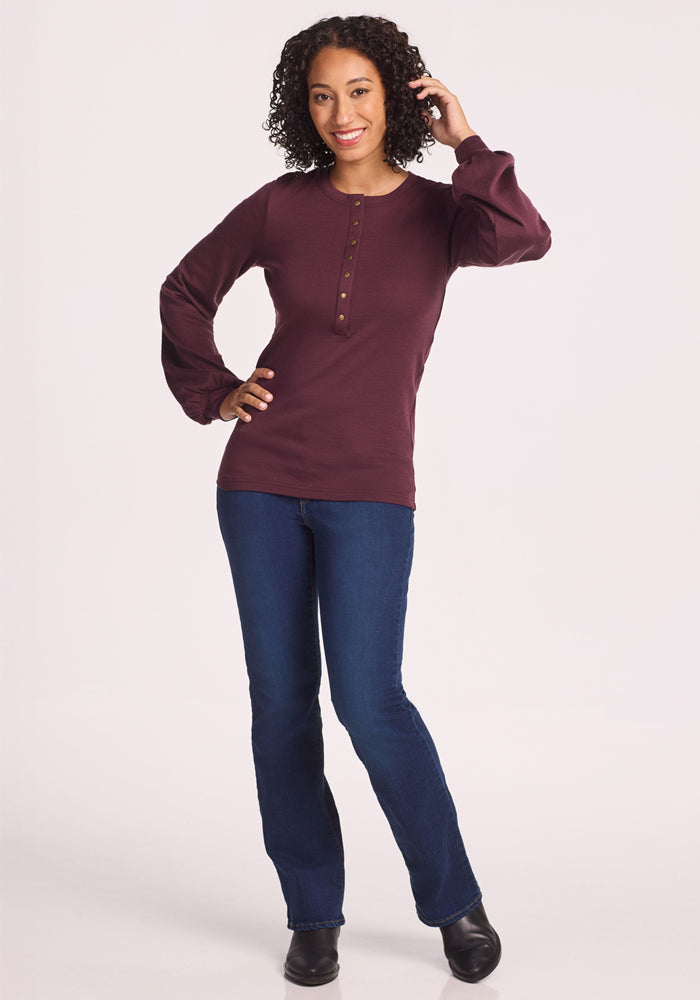 A woman with curly hair wears the Woolx Charlotte Puff Sleeve Top in Chocolate Fig, blue jeans, and black shoes. She smiles, one hand touching her hair and the other on her hip, against a plain light background.