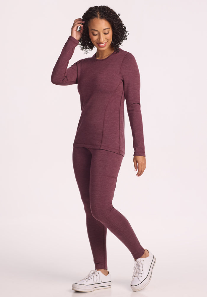 A person with curly hair smiles while wearing the Woolx Elsa Long Sleeve in Grape Harvest Melange, paired with matching leggings and white sneakers. They stand against a light background, touching their hair with one hand.