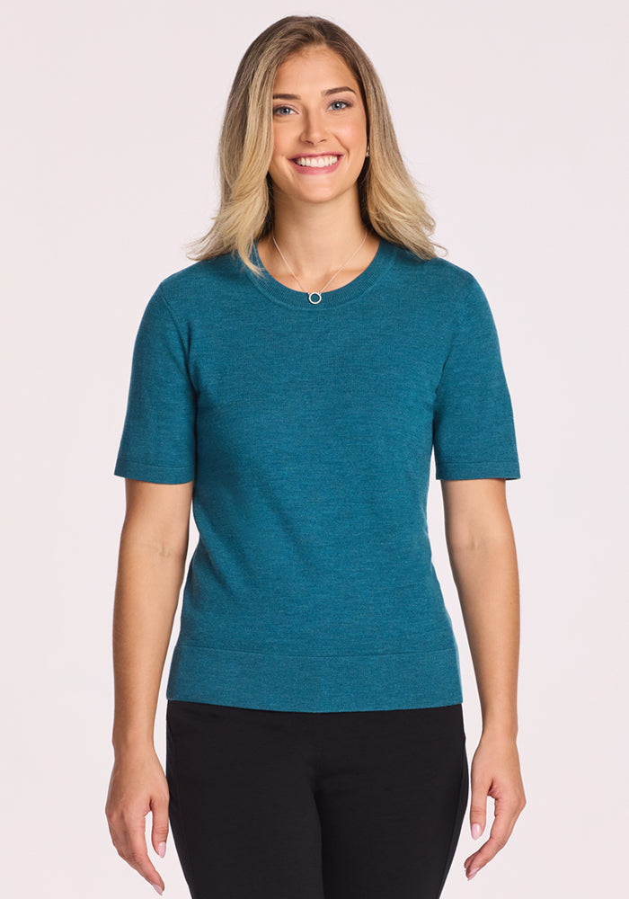A smiling woman with shoulder-length blonde hair wears the Woolx Anna Crew Sweater - Harbor Teal, a stylish women's short-sleeve sweater, paired with black pants against a plain light background. 