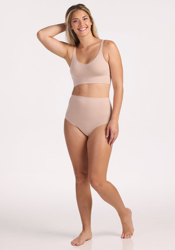 A smiling woman stands barefoot against a white background, wearing the Woolx Mabel Full Coverage Underwear in Sand. One hand rests on her head as she exudes seamless comfort and confidence in this high-waisted, light beige style.