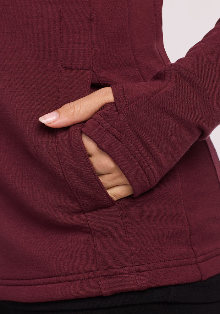 A close-up of a hand with light pink nails sliding into the pocket of a maroon Woolx Zoey Zip Up Hoodie in Toasted Garnet, highlighting the visible stitching and seam details.