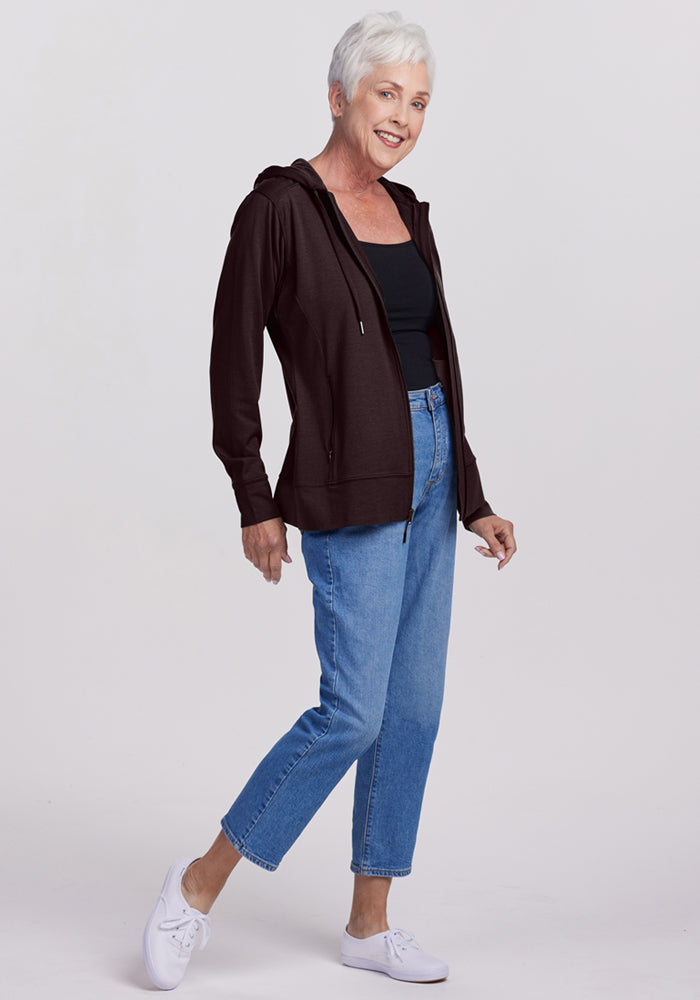 Older woman with short gray hair wears the Woolx Ryann Hoodie in French Roast over a black top, blue jeans, and white sneakers. She smiles while standing against a plain light background.