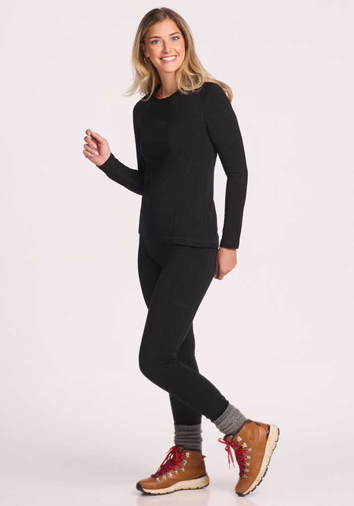 A smiling woman wears the Woolx Elsa Long Sleeve - Black base layer top and leggings, paired with gray socks and tan hiking boots with red laces, standing confidently against a plain light background.