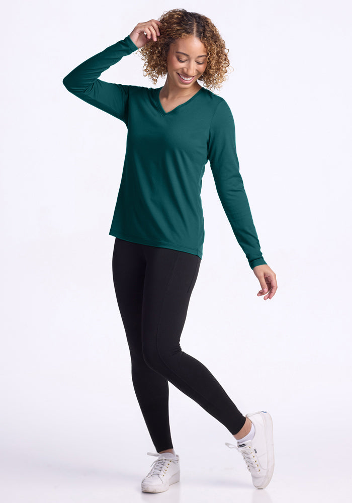 A smiling woman with curly hair wears the Woolx Millie Long Sleeve in Riverbank Green, paired with black leggings and white sneakers. She stands on one foot, hand in her hair, against a plain white background.