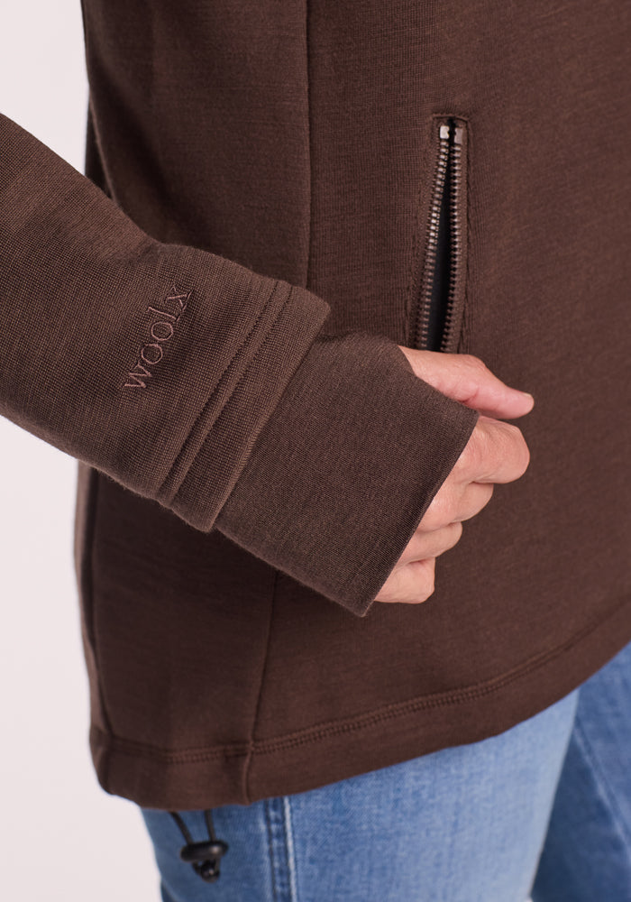 A woman wears the Woolx Cubby Hooded Sweatshirt in Bracken, featuring thumb holes, an embroidered "wool" logo, a side zippered pocket, and pairs it with blue jeans for a warm fleece look.