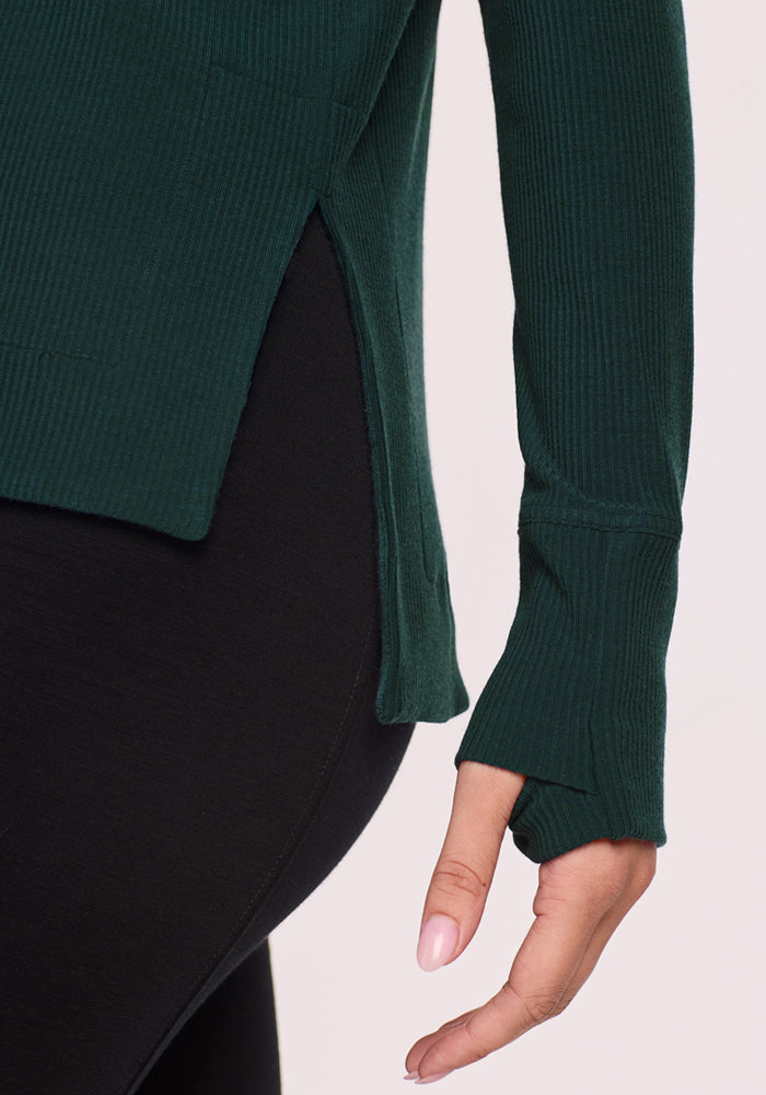 A close-up shows the arm, hand, and lower torso of a person wearing the Woolx Ainsley Sweater in Deep Timber, a women’s ribbed style with side slit and thumbhole, paired with black fitted pants.