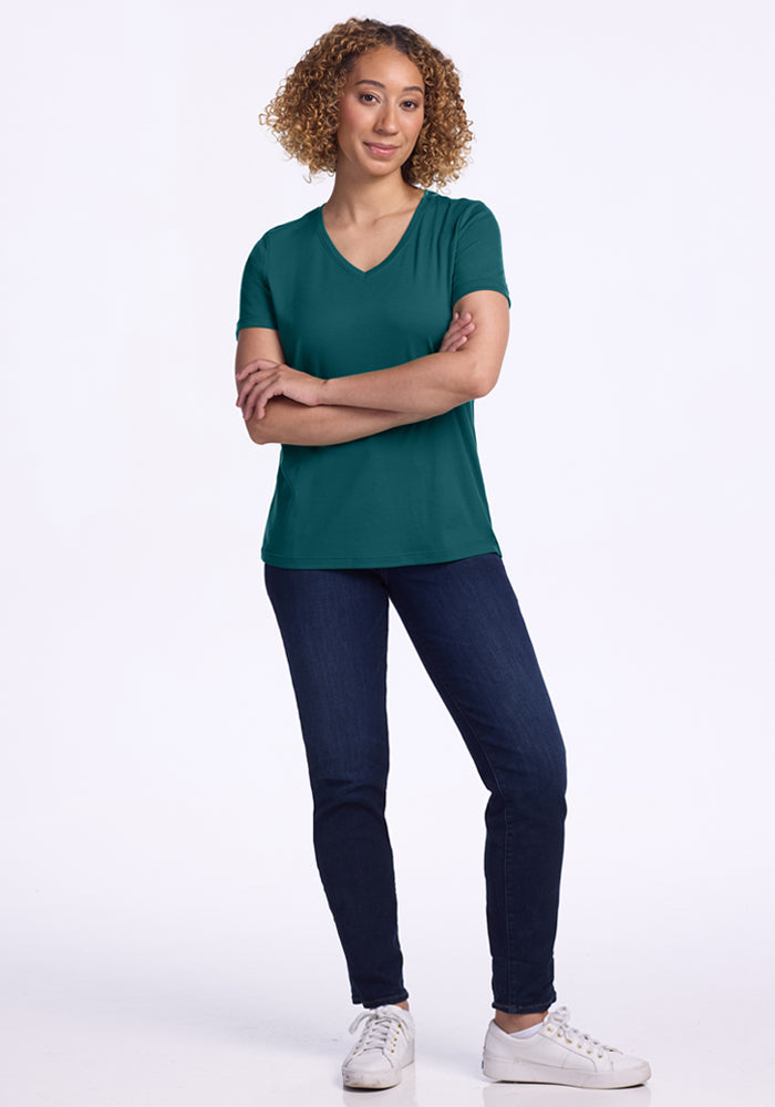 A woman with curly hair stands against a white background, wearing the Woolx Junie V Neck in Riverbank Green, paired with dark blue jeans and white sneakers. Her arms are loosely crossed as she looks at the camera with a slight smile.