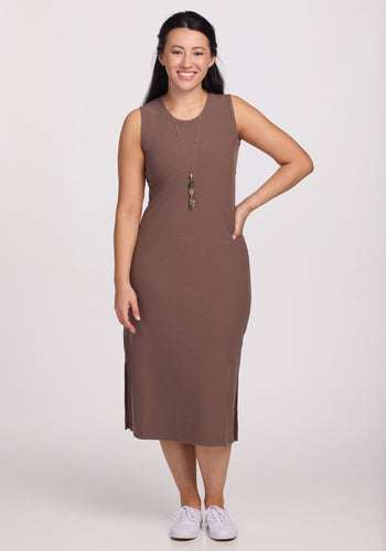 A woman in the Woolx Iris Ribbed Dress - Toasted Almond, paired with white sneakers, stands smiling against a plain background. Her long hair complements her long necklace. The odor-resistant and wrinkle-resistant fabric keeps her looking fresh throughout the day.