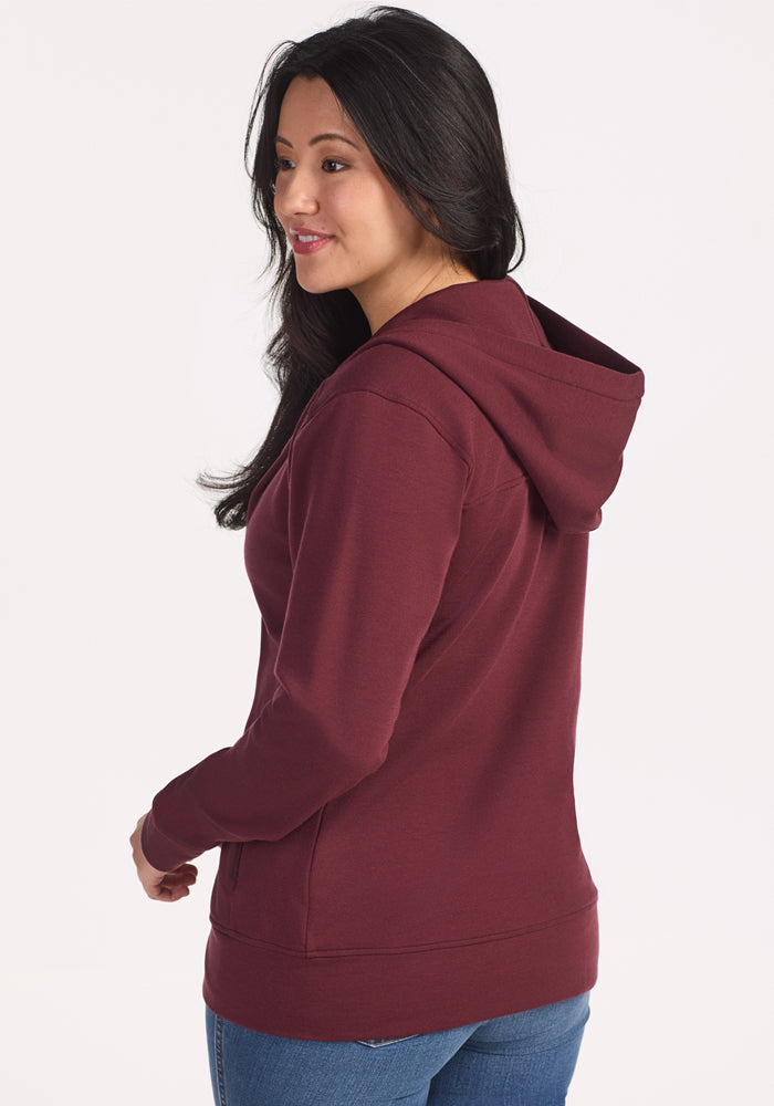 A woman with long dark hair, wearing the Woolx Darby Zip Up Hoodie in Toasted Garnet and blue jeans, stands facing slightly away from the camera, smiling as she looks to the side.