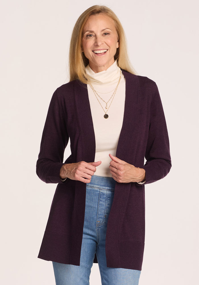 A smiling woman with straight blonde hair wears the Woolx Macie Sweater in Deep Plum over a white turtleneck, paired with layered gold necklaces and blue jeans, standing against a plain light background. 
