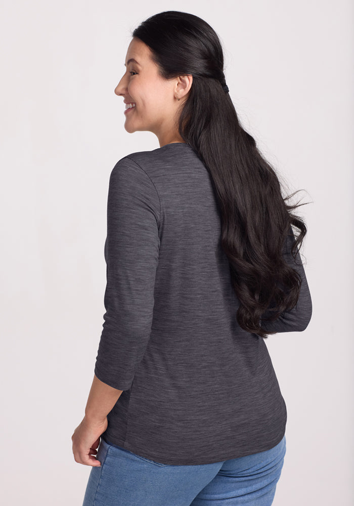 A woman with long dark hair, styled half-up, stands smiling and facing away. She wears the Woolx Jenny 3/4 Sleeve Crew Neck in Cinder Melange and blue jeans against a plain light background.