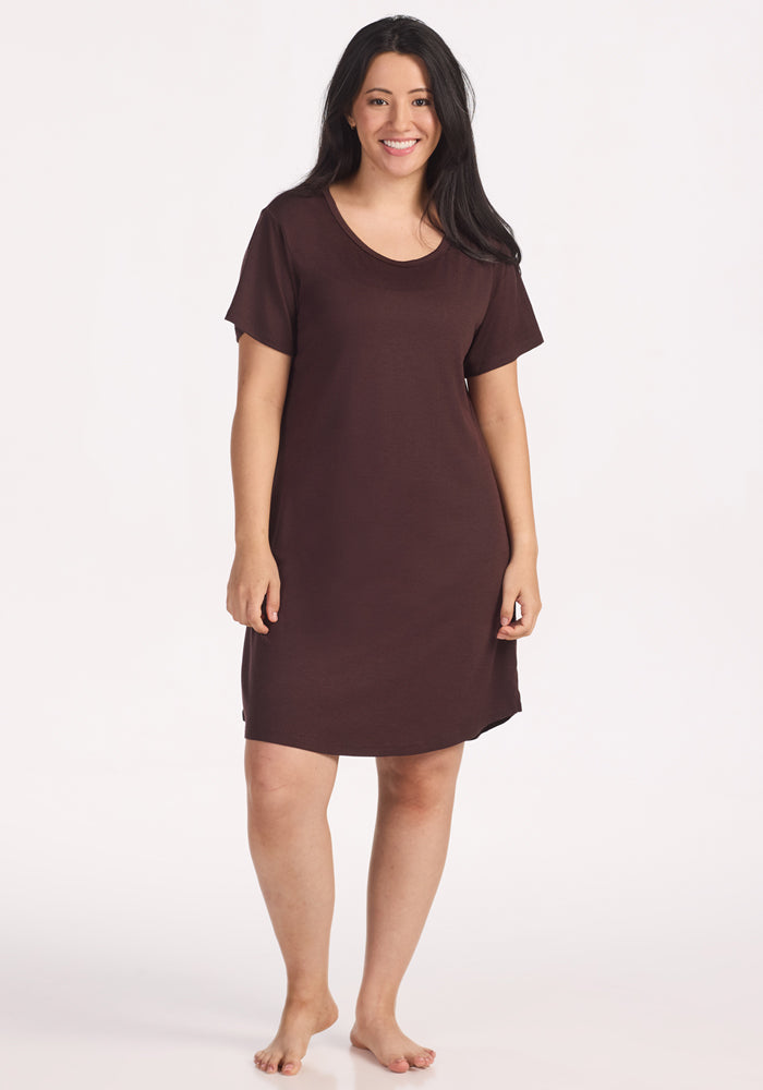 A woman with long dark hair smiles barefoot in the Woolx Desi Nightgown - Chocolate Plum, a short-sleeve, loose-fitting merino wool dress that falls just above her knees, standing against a plain white background. 