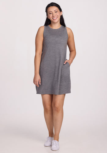 Against a white background, a person smiles in the Clara Dress - Graphite Heather from Woolx, crafted from temperature-regulating Merino wool, paired with white sneakers and one hand in their pocket. | Sarah is 5’8”, wearing a size S