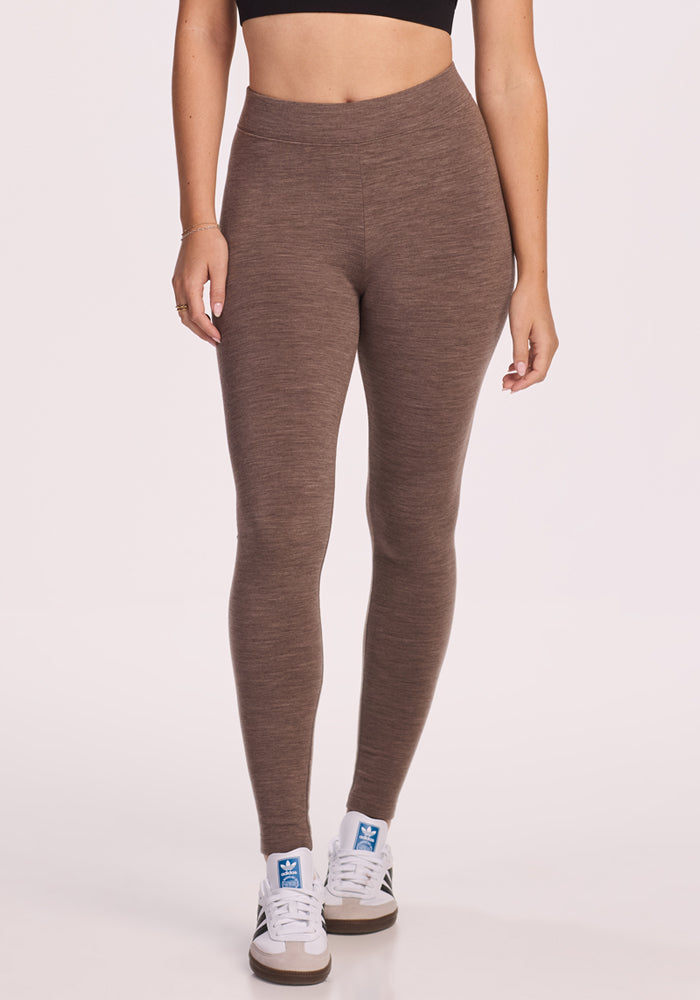 A person stands against a plain, light background wearing Woolx Stella Petite leggings in Simply Taupe, a black crop top, and white Adidas sneakers. Photo is cropped from the shoulders down.