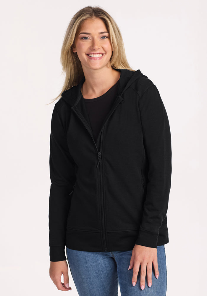 A smiling woman with long blonde hair wears the Woolx Darby Zip Up Hoodie in black over a black top and blue jeans, standing against a plain light background. 