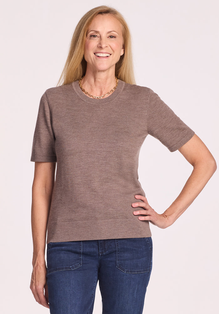 A woman with long blonde hair smiles, standing with one hand on her hip. She wears the Woolx Anna Crew Sweater in Simply Taupe, blue jeans, and a gold chain necklace against a plain light background. 