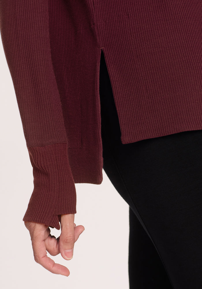 A close-up of a person wearing the Woolx Ainsley Sweater in Toasted Garnet, featuring premium merino wool, ribbed texture, side slit, and thumbhole cuffs—styled with black pants for a classic women's sweater look. Only the hand, arm, and torso are visible.