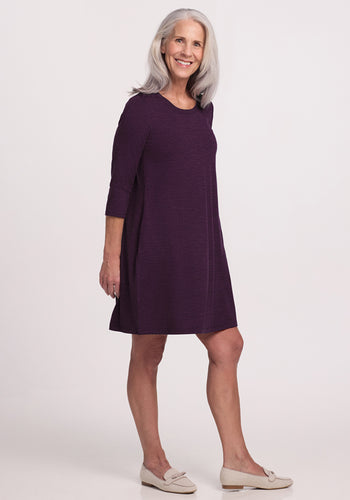 A smiling older woman with long gray hair wears the Woolx Rissa A Line Dress in Deep Plum—knee-length with long sleeves—paired with beige slip-on shoes, perfect for fall and winter layering, standing against a plain white background. | Tam is 5’8.5”, wearing a size S