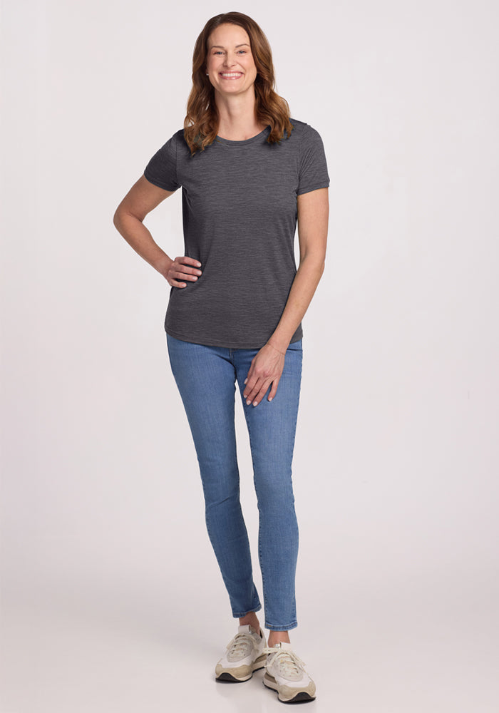 A person smiles against a plain light background, wearing the Woolx Liza Crew Neck Tee in Cinder Melange, paired with blue jeans and white sneakers, with one hand on their hip and the other relaxed by their side.